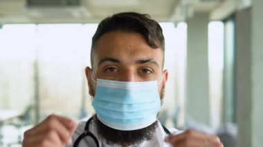 Tıbbi maskeli ve steteskoplu beyaz önlüklü Arap doktor stajyer doktor klinikte duruyor.