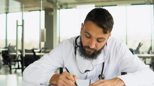 身穿白色工作服的阿拉伯年轻医生坐在工作场所 在纸质杂志上写着便条 — 图库视频影像