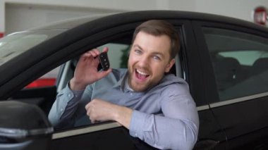 Neşeli genç adam yeni otomobil salonunda oturuyor ve anahtarlarını gösteriyor. Mutlu araba sahibi..