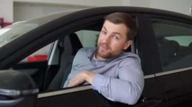 Neşeli genç adam yeni otomobil salonunda oturuyor ve anahtarlarını gösteriyor. Mutlu araba sahibi.