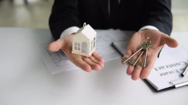 Kapalı görüşlü emlakçı elinde ev anahtarı ve beyaz ev modeli tutuyor.