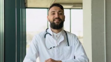 Beyaz üniformalı gülümseyen Arap erkek doktor hastanede iyi hizmet veriyor. Klinikteki mutlu pratisyen hekimin ya da çocuk doktorunun portresi. Sağlık hizmetleri kavramı.