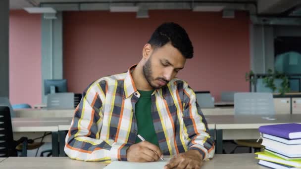 准备考试 有思想的印度学生坐在图书馆书桌边 一边在记事本上写东西 一边看书 — 图库视频影像
