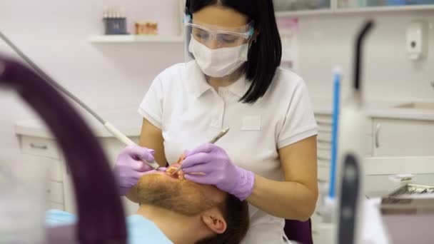 顔のマスクと眼鏡の女性歯科医は 男性患者のための治療を行い 歯のツールを保持し ゴム手袋を着用 血液学 歯科学 現代の歯科クリニックのコンセプト — ストック動画