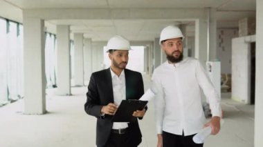 Hintli ve Arap inşaatçılar miğferli işçiler güneş ışığı araştırmasında inşaat üzerinde yürüyorlar şehir merkezinde gökdelenle buluşuyorlar.