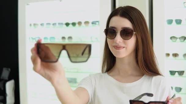 在眼镜店里表现出快乐而犹豫的女人的形象 做决定 拿着时髦的太阳镜 选择她应该买的东西 哪一个更适合我 买两双的折扣很大 — 图库视频影像