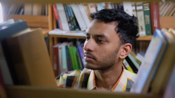 聪明迷人的印度男生或自由职业者 穿着休闲装 站在书架上的图书馆里 自己选一本书 从书架上取下 — 图库视频影像