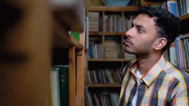 聪明迷人的印度男生或自由职业者 穿着休闲装 站在书架上的图书馆里 自己选一本书 从书架上取下 — 图库视频影像