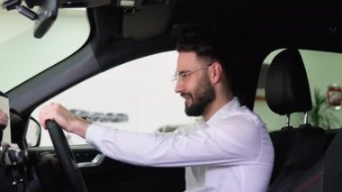 Happy man sits in new electric car in shop dealership and celebrate purchase of new vehicle. Buy car