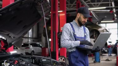 Üniformalı deneyimli tamirci otomobil servisinde araba tamir ederken bir dizüstü bilgisayar kullanıyor.