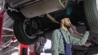 Profesyonel Tamirci Eldiven Giyiyor ve Arabanın Altında Ratchet Kullanıyor. Bir araba servisinde çalışan yakışıklı bir tamircinin portresi çekiliyor. Modern Temiz Atölye.