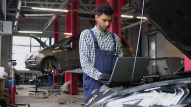 Üniformalı atölyedeki Hintli adam bozuk arabayı tamir etmek için dizüstü bilgisayar kullanıyor.