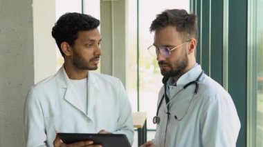Tıbbi ekip iki çok ırklı erkek doktor beyaz önlüklü konuşmalar yapar, dijital tablet kullanır. Tıp konsepti ortaklığı, sağlık teknolojisi.