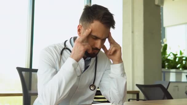 疲惫的医生穿着白色制服 一个人坐在医院的办公桌前头疼 可怜的年轻人对自己的医疗失败感到绝望和内疚 — 图库视频影像