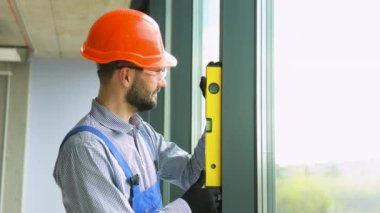 Üniformalı bir tamirci yeni ofise PVC pencereler takıyor. Bir adam pvc pencerelerinin dikey sapmasını ölçer