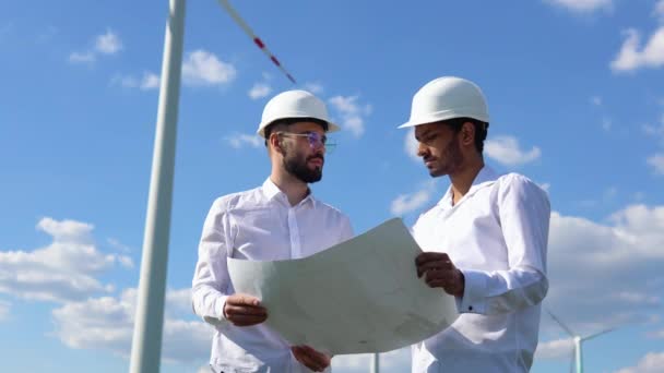 两名多族裔工程师讨论了一个风力发电厂的蓝图 该发电厂位于一个通过转换动能提供替代电力的涡轮机前面 — 图库视频影像