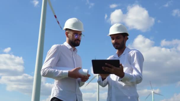 白人検査官との会談中にタブレットを使用してヘルメットでインドのエンジニア 風車で農場に立っている間に緑のエネルギーについて議論する2人のパートナー — ストック動画