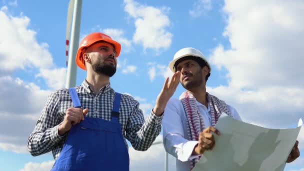 两名多族裔工程师讨论了一个风力发电厂的蓝图 该发电厂位于一个通过转换动能提供替代电力的涡轮机前面 — 图库视频影像