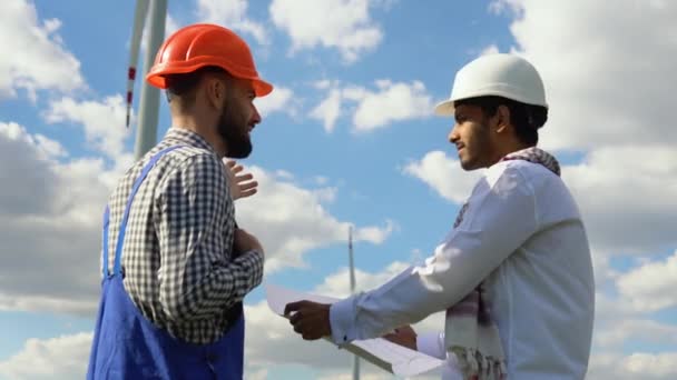 Dwóch Wielonarodowych Inżynierów Omawiających Plan Budowy Farmy Wiatrowej Przed Turbiną — Wideo stockowe