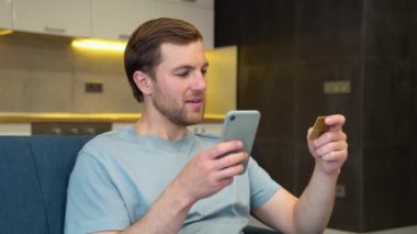 İnternet Mobil Alışveriş Konsepti. Pozitif Ortadoğulu erkek cep telefonu kullanıyor. İnternetten bir şeyler alıyor. Kanepede oturuyor, web mağazasından hediyeler alıyor.