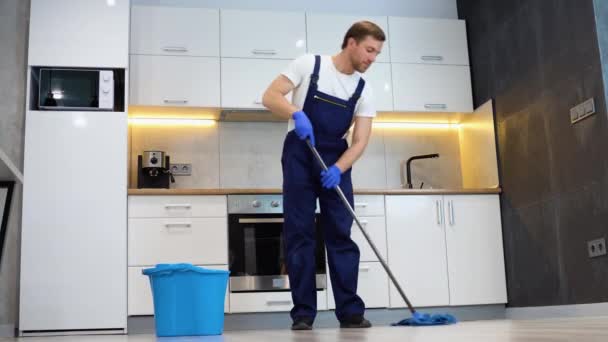 作为一名职业清洁工的男人 穿着蓝色制服 在厨房里用拖把和桶擦洗地板 — 图库视频影像