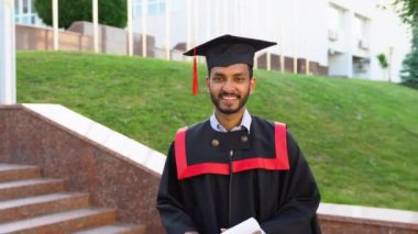Mezuniyet elbisesi ve şapkasıyla kampüste diplomasıyla yürüyen gülümseyen Hintli öğrenci..