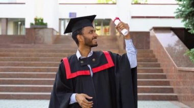 Mezuniyet kostümlü neşeli Hintli adam diplomasını gösteriyor ve kameraya gülümsüyor..