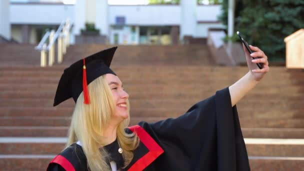 卒業証書付きの自撮り 卒業式で幸せそうな金髪の女子大生 魅力的な女性は スマートフォンを見て セルフィーを作る — ストック動画