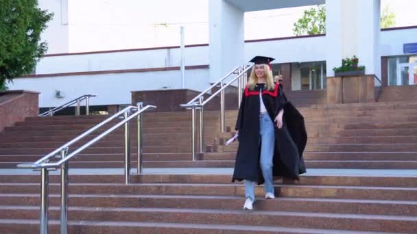 캠퍼스에서 가운을 모자를 졸업장을 가지고 다니는 금발의 여성에게 미소짓는 — 비디오