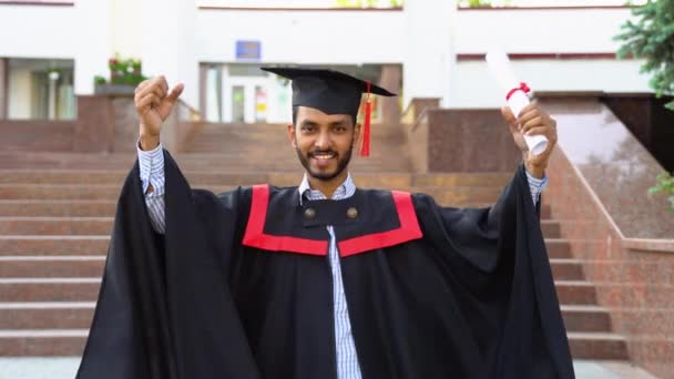 モルタルボードの幸せなインドの男性大学院生とキャンパス内の卒業証書と学士号のガウン 教育や卒業や人々の概念は — ストック動画