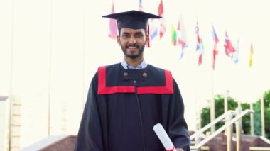 Mezuniyet kostümlü neşeli Hintli adam diplomasını gösteriyor ve kameraya gülümsüyor..