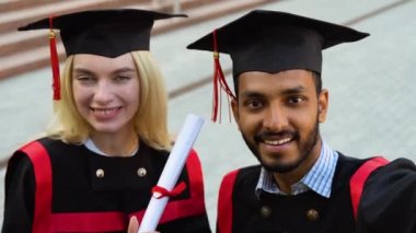 Mezuniyet elbiseleri ve şapkalarıyla birlikte selfie çeken, üniversite kampüsünde poz veren mezuniyetin tadını çıkaran ve kutlayan mutlu çok ırklı çift öğrenciler..
