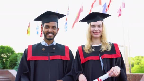 幸せな卒業生の肖像画 卒業キャップとガウンを着た2人の友人が大学の建物の外に立って卒業証書の巻物を持って笑顔で — ストック動画