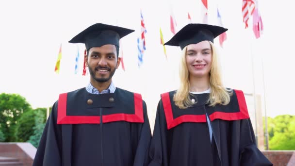 幸せな多民族卒業生の肖像画 卒業キャップとガウンを着た2人の友人が大学の建物の外に立って卒業証書の巻物を持って笑顔で — ストック動画