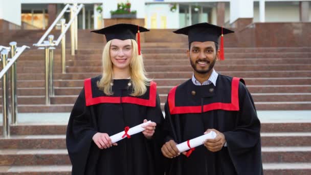 幸せな多民族卒業生の肖像画 卒業キャップとガウンを着た2人の友人が大学の建物の外に立って卒業証書の巻物を持って笑顔で — ストック動画