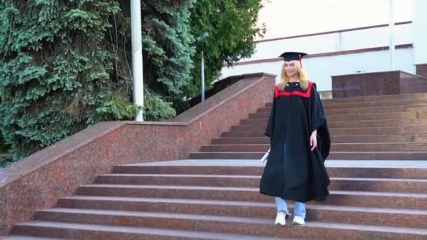 笑顔大学院ブロンド女の子で卒業生ガウンと帽子歩くととともにディプロマでキャンパス — ストック動画