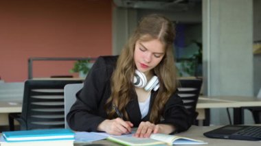 Konsantre genç kız öğrenci tahta masada dizüstü bilgisayarla oturuyor ve kütüphanede sınava hazırlanırken notlar yazıyor..