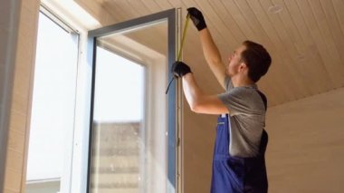 Üniformalı servis elemanı, ahşap prefabrik evde ölçü bandıyla PVC penceresi kuruyor.