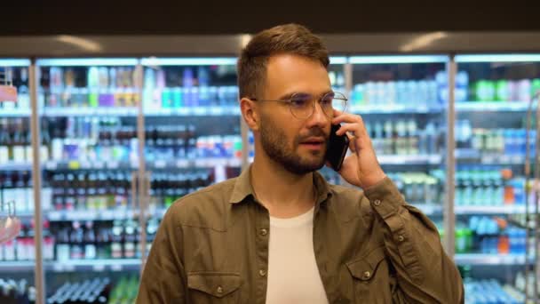 Молодой Человек Входит Супермаркет Делает Покупки Выбирает Продукты Говорит Телефону — стоковое видео