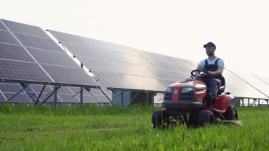 Bir adam güneş panellerinin yakınındaki çimleri biçiyor. Yeşil enerji.