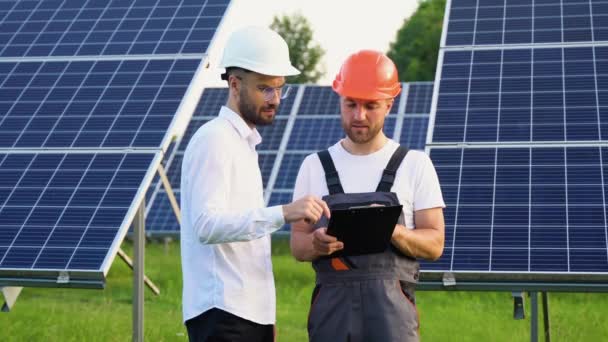 大きな太陽電池パネルを背景に青い印刷物を持ちながら話をする2人のエンジニア — ストック動画