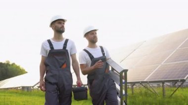 Güneş enerjisi santrali. İki mühendis koruyucu iş kıyafetleri içinde. Yürüyen ve fotovoltaik panelleri inceliyorlar. Alternatif enerji kavramı ve onun bakımı.
