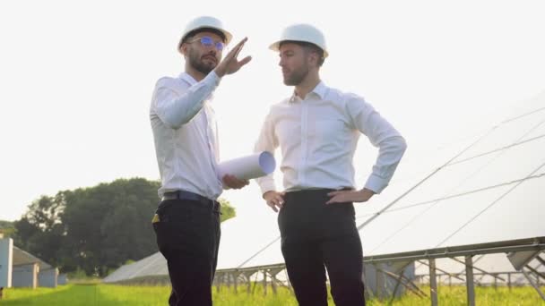 ソーラーファームでのプロジェクトについて話し合う2人のハードヘルメットの男性 技術図面通信のスクロールを持つ男性エンジニア 緑のエネルギーの概念 — ストック動画