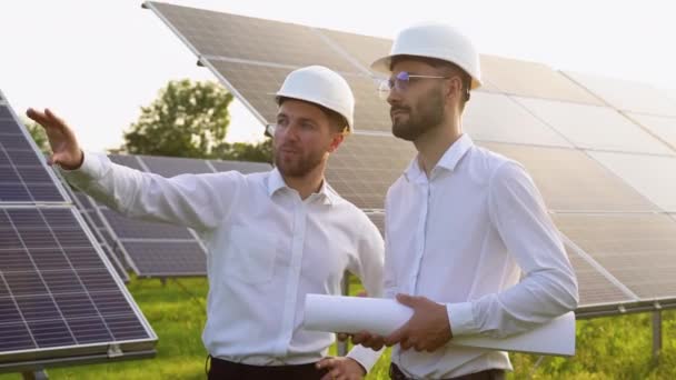 两个戴硬盔的男人在太阳能农场讨论项目 拥有工程图纸交流卷轴的男性工程师 绿色能源的概念 — 图库视频影像