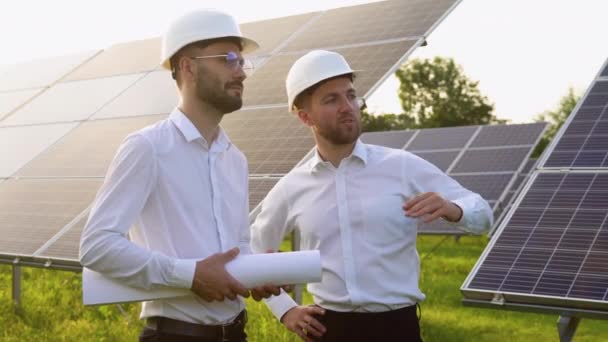 两个戴硬盔的男人在太阳能农场讨论项目 拥有工程图纸交流卷轴的男性工程师 绿色能源的概念 — 图库视频影像