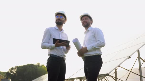 两个戴硬盔的男人在太阳能农场讨论项目 拥有石碑和工程绘图卷轴的男性工程师进行沟通 绿色能源的概念 — 图库视频影像