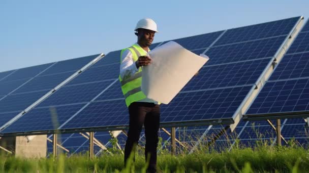 身穿白色安全帽的年轻黑人工程师手握项目文件计划 对太阳能发电厂的光伏电池板进行了验证 — 图库视频影像