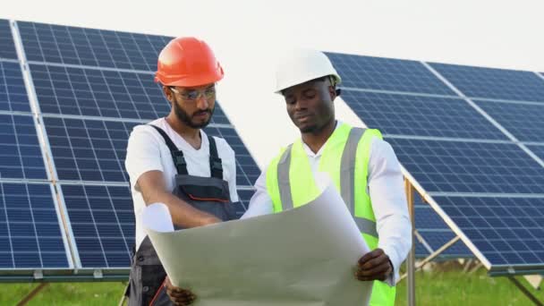 黑人工程师和印第安工人彼此交谈 同时拿着一个蓝色的蓝图 背景上有一个巨大的太阳能电池板 — 图库视频影像