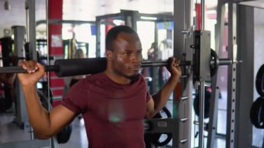 Siyah vücut geliştirici omuzunda halterle çömeliyor. Afrikalı adam spor salonunda ağırlık kaldırıyor. Güçlü kaslı adam spor salonunda ağırlık halterini kaldırmaya çalışıyor..