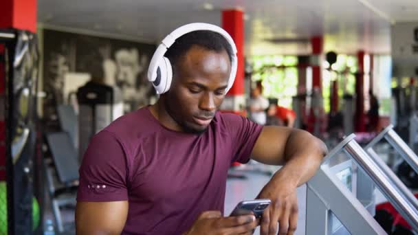 ワイヤレスヘッドセットとタオルを備えたリラックスしたアフリカ系アメリカ人のスポーツマンは ワークアウト ジムインテリアのための新しい音楽アプリケーションを使用して 携帯電話で音楽を聴きます — ストック動画